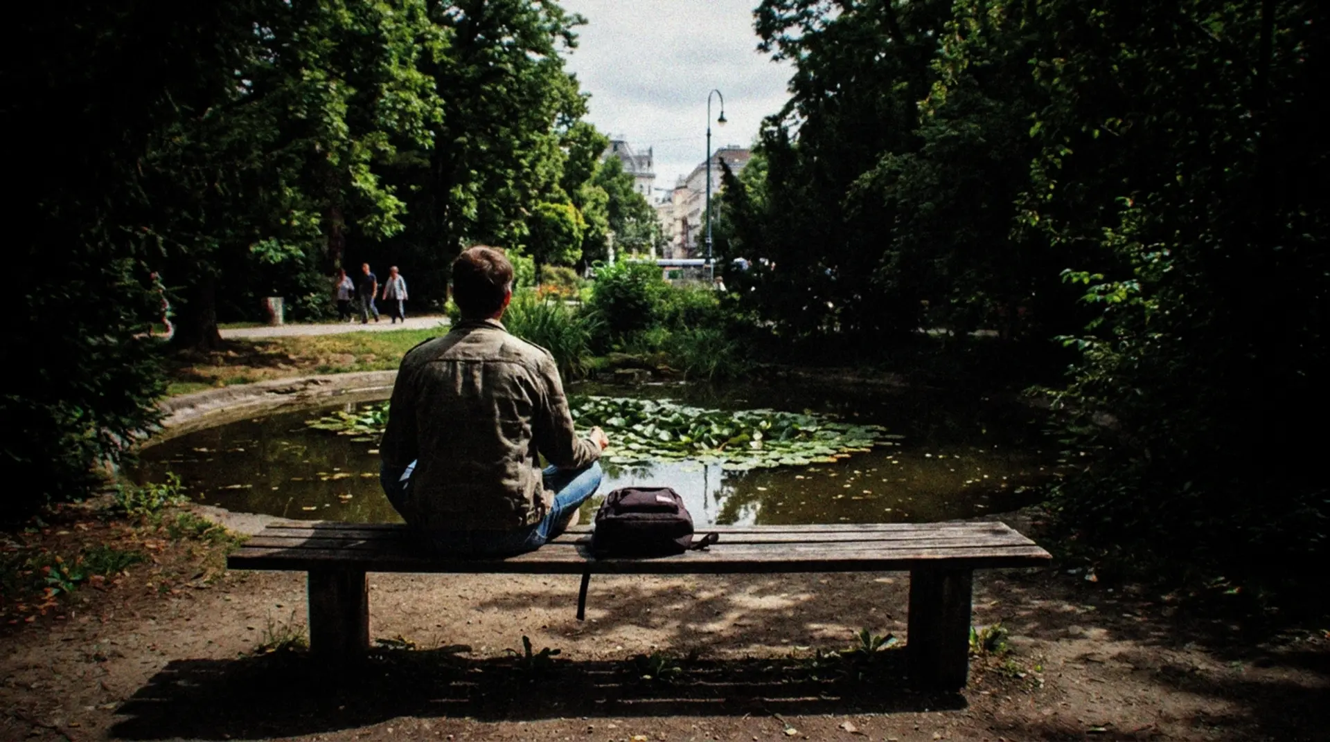 A person sitting in meditation pose on a wooden bench in a secluded corner of Vienna's Stadtpark, surrounded by lush green trees and flowering plants. Their back is to the camera as they face a small ornamental pond with water lilies. Morning light filters through the leaves, creating dappled shadows on the ground. A few other park visitors can be seen in the far distance walking along paths. The scene captures the contrast between Vienna's urban environment visible in the background through gaps in the trees and this peaceful natural sanctuary in the foreground. The meditator wears casual clothing and has a small backpack resting beside them on the bench.
