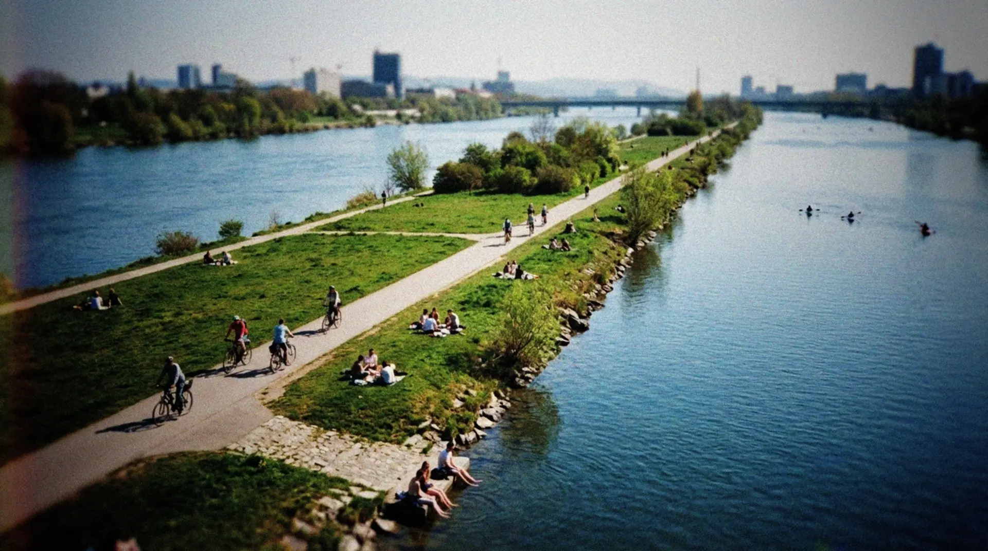 A candid documentary-style photo of Danube Island in Vienna on a sunny spring day, captured from a slightly elevated perspective. The narrow strip of land stretches through the frame with the blue Danube River visible on both sides. People can be seen enjoying recreational activities - cyclists riding along pathways, small groups picnicking on grassy areas, and a few individuals relaxing on the shoreline with their feet in the water. In the distance, kayakers paddle along the calm water. The Vienna skyline appears subtly in the background, showing the contrast between urban life and this natural recreational space. The photo has a casual, spontaneous quality as if taken with a smartphone during a weekend outing, with natural lighting and realistic colors.
