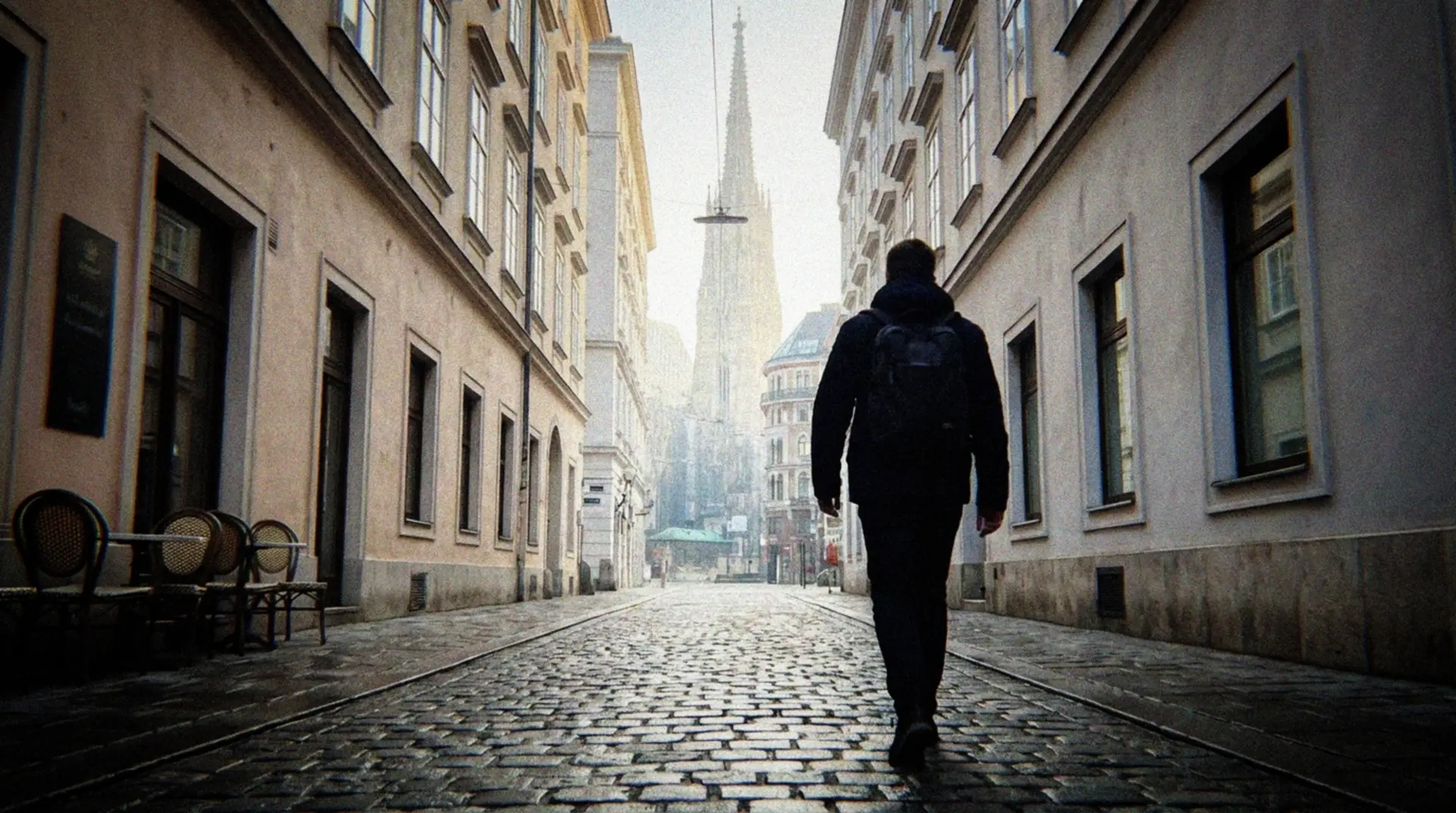 A solitary figure walking thoughtfully along a quiet cobblestone street in Vienna's old town at early dawn, photographed from behind. The morning light is soft and natural, illuminating the historic facades of traditional Viennese buildings with their distinctive architecture. The street is nearly empty, with just hints of the city awakening - perhaps a distant café worker setting up tables or a street cleaner. Subtle morning mist hangs in the air, creating a contemplative atmosphere. The walker wears casual, comfortable clothing and appears to be moving at a slow, deliberate pace, suggesting mindfulness rather than hurried tourism. The perspective captures both the walker and the beautiful architectural details of Vienna, with perhaps Stephansdom cathedral visible in the distance. The scene has a casual, unstaged quality as if captured spontaneously with a smartphone during an early morning walk.