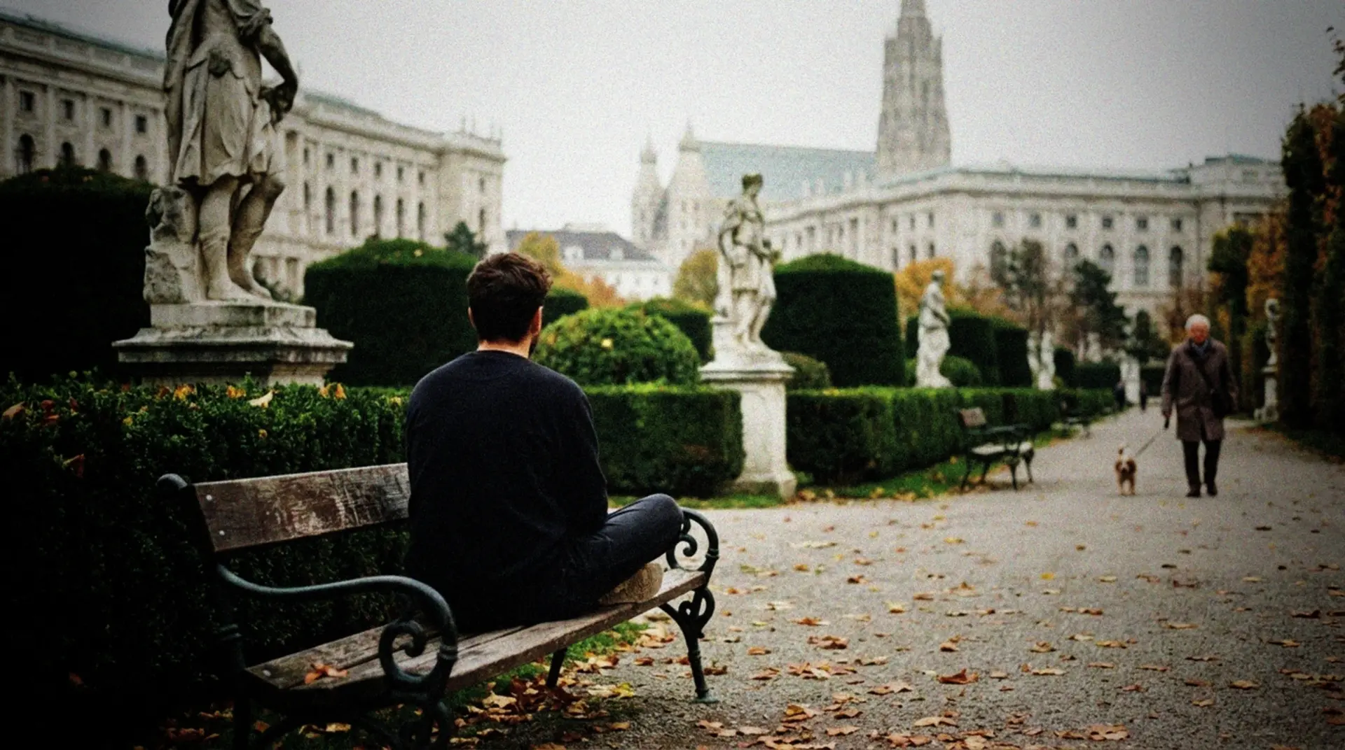 A solitary person sitting cross-legged in meditation on a wooden bench in one of Vienna's peaceful public gardens, surrounded by neatly trimmed hedges and classical statues. The historic architecture of Vienna is visible in the soft background, with the distinct spires of St. Stephen's Cathedral barely visible in the distance. The person is seen from behind, dressed in casual clothing, their posture relaxed but attentive. A few autumn leaves scatter the ground, and an elderly local walks a small dog along a distant pathway. The natural daylight creates a calm atmosphere without being overly stylized - just an honest snapshot of a quiet moment in an otherwise bustling European capital. The contrast between the stillness of the meditating figure and the grand imperial surroundings captures the essence of finding mindfulness in an urban setting.