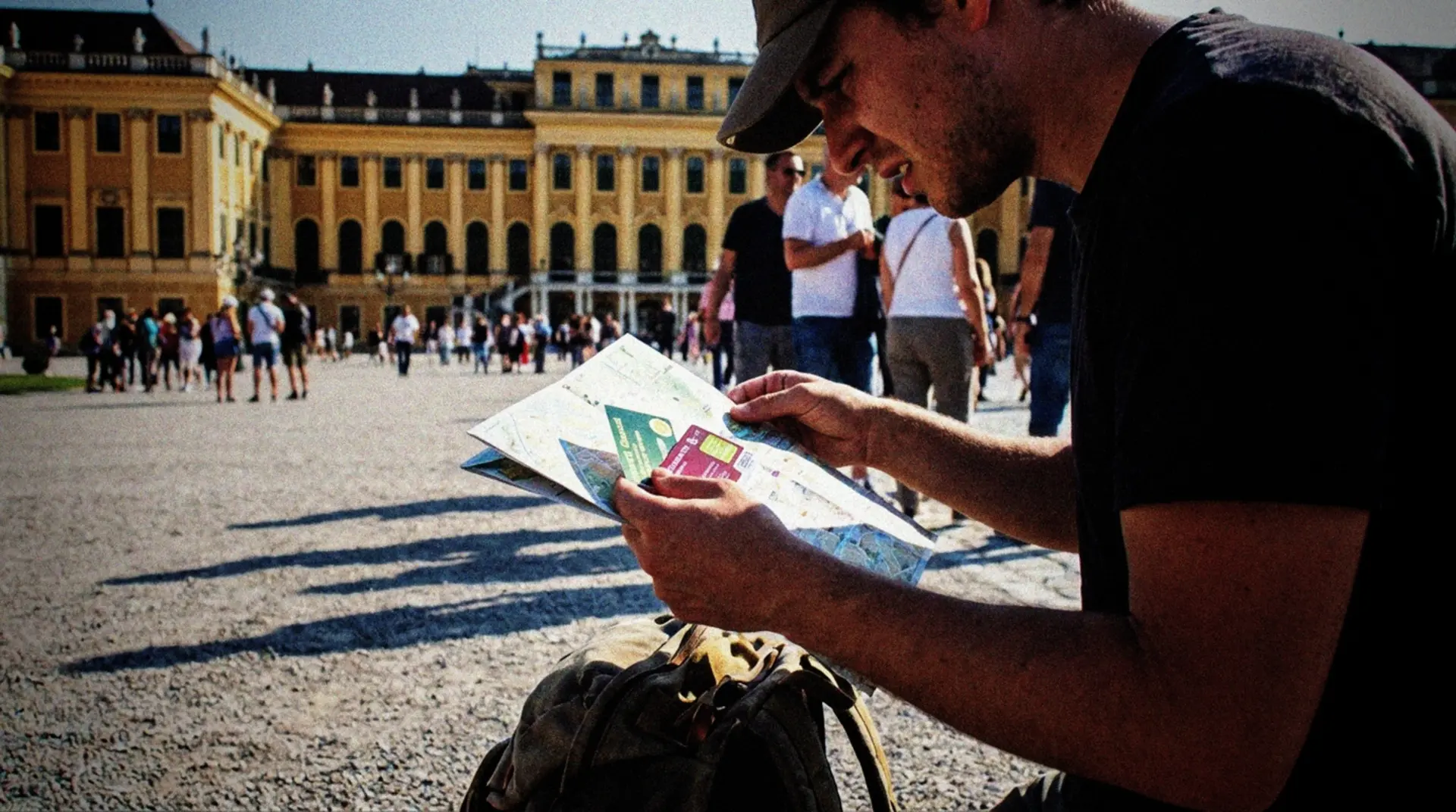 A tourist standing at the entrance to Schönbrunn Palace in Vienna, looking at a city pass and a map. Shot from a side angle with the tourist's face partially obscured, they appear slightly confused while comparing what appears to be two different types of tourist cards. In the background, the magnificent yellow facade of the palace is visible with other tourists walking around. A small backpack rests at the person's feet, and the bright daylight creates natural shadows across the scene. The casual, documentary-style composition captures an authentic moment of travel decision-making in one of Vienna's most iconic locations.